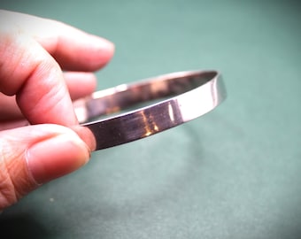 May include: A silver-colored, flat bangle bracelet. The bracelet is held by a hand against a green background. The bangle has a simple, minimalist design, with a smooth, reflective surface. The bracelet appears to be made of metal.