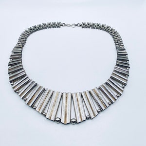 Puede incluir: Un collar de plata con un diseño geométrico. El collar está hecho de pequeñas piezas rectangulares de metal que están conectadas entre sí. El collar tiene un cierre en la parte posterior.