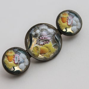 Könnte beinhalten: Eine Vintage-Brosche mit drei runden Glascabochons, die handbemalte gelbe und weiße Narzissen mit grünen Blättern zeigen. Die Brosche ist aus Metall mit einer goldenen Oberfläche.