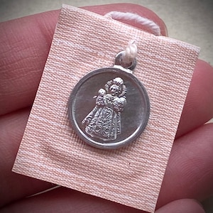 Peut inclure: Un charme en argent représentant une image détaillée d'une figure religieuse, probablement une sainte, tenant un enfant. Le charme est sur un fond de tissu blanc.