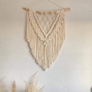 Könnte beinhalten: Cremefarbener Makramee-Wandbehang mit geometrischem Design. Das Stück ist an einem natürlichen Holzdübel aufgehängt und weist komplizierte Knotenmuster auf, die eine strukturierte, böhmische Ästhetik erzeugen. Der Boden verjüngt sich zu einem Punkt.