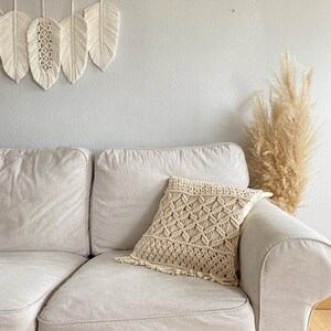 May include: A white couch with a cream-colored throw pillow with a macrame design. The couch is in front of a white wall with a macrame wall hanging.