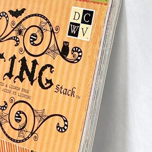 May include: A paper stack pad with an orange striped cover featuring black illustrations of bats, owls, and spiderwebs. The text reads DCWV, ING, Stack, and Acid & Lignin Free. The top edge has a green border with blue stars.