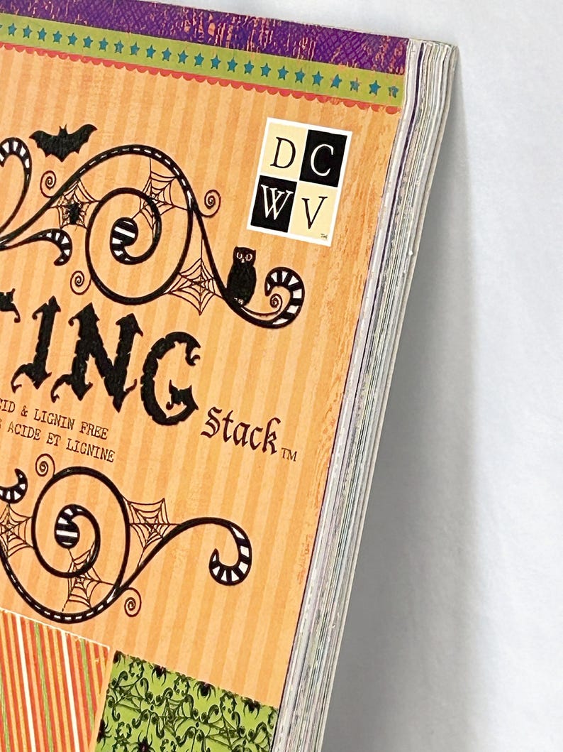 May include: A paper stack pad with an orange striped cover featuring black illustrations of bats, owls, and spiderwebs. The text reads DCWV, ING, Stack, and Acid & Lignin Free. The top edge has a green border with blue stars.