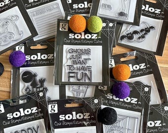 Op de afbeelding: Een verzameling Studio G Soloz transparante stempelsets met Halloween-thema's zoals geesten, vleermuizen, spinnen en spookhuizen. Elke verpakking toont de tekst GHOULS JUST WANT TO HAVE FUN of andere zinnen, geaccentueerd door kleine vilten pompons.