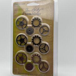 May include: A package of 12 metal gears in various sizes and shapes. The gears are in a clear plastic package with a brown and green background. The package has the text "SPROCKET GEARS 12 GEARS" and "Jim Holtz idea-ology" on it.