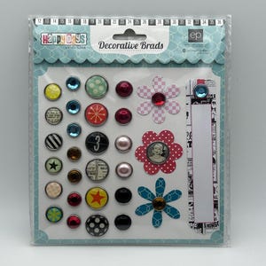 May include: A sheet of decorative brads with various designs and colors, including stars, flowers, numbers, and geometric shapes. The brads are packaged in a clear plastic sheet with the text "Happy Days" and "Decorative Brads" printed on it.
