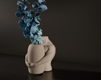 May include: A beige ceramic vase in the shape of a hugging figure, holding a smaller vase filled with blue-green leafy stems. The vase is on a dark surface with a dark background.
