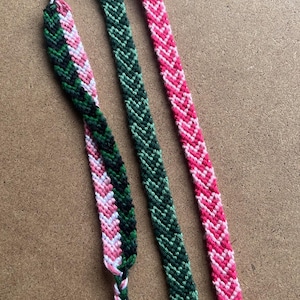 May include: Three friendship bracelets made with colourful embroidery floss. One bracelet is pink and white, one is green and black, and one is green and white.