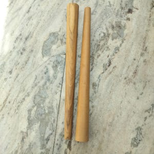 Könnte beinhalten: Zwei Holzdübel, einer zylindrisch und der andere konisch, werden vor einer hellen, gesprenkelten Oberfläche gezeigt. Die Dübel sind aus hellem Holz gefertigt und zeigen die natürliche Maserung. Der zylindrische Dübel hat einen Durchmesser von etwa 2,5 cm.