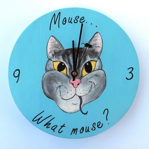 May include: A blue round clock with a gray cat face painted on it. The cat has yellow eyes and a pink nose. The clock hands are black and the clock face says "Mouse... What mouse?" and has the numbers 3 and 9.