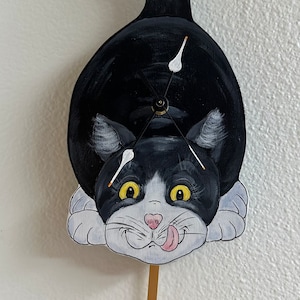 May include: A whimsical, hand-painted cat clock with a black and white body, yellow eyes, and a pink tongue sticking out. The clock has a pendulum in the shape of an orange fish. The clock hands are white.