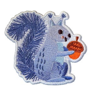 May include: An embroidered patch featuring a light blue squirrel with a large, bushy tail and tufted ears. The squirrel is holding a bright orange acorn with a brown cap. The entire design is outlined with a white stitched border on a white background.