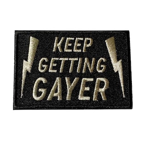 May include: Black embroidered patch with the text "KEEP GETTING GAYER" in a bold, stylized font. Two gold lightning bolts flank the text. The patch is rectangular and has a black border.