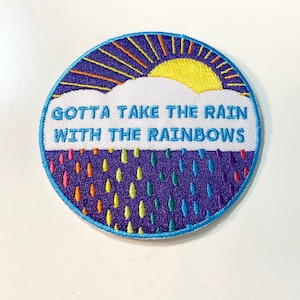 May include: A round, purple and blue embroidered patch with a white cloud and yellow sun. The text reads "Gotta take the rain with the rainbows".