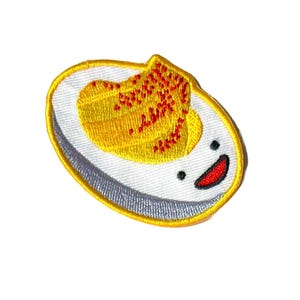 May include: A white embroidered patch with a yellow egg yolk with red sprinkles on top. The egg white is a cartoon face with a smiling mouth and two black eyes.
