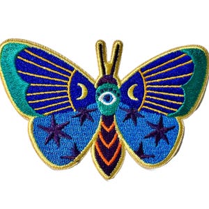 May include: Embroidered iron-on patch featuring a blue butterfly with a green eye, gold accents, and crescent moons on the wings.