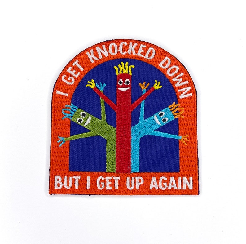 以下が含まれることがあります： オレンジ色の刺繍パッチに「I get knocked down but I get up again」の文字が書かれています。パッチには、笑顔の顔をした風船人形のキャラクターが3人描かれています。