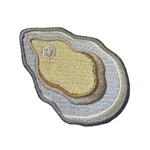 May include: An embroidered patch shaped like an oyster shell. The center is a textured pale yellow, surrounded by layers of light gray and white thread. The outer edge is defined by a dark gray stitched border, creating a distinct shell silhouette.