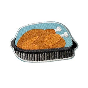 May include: Embroidered patch featuring a roasted turkey in a black roasting pan, set against a light blue background. The turkey is golden brown with white bone details. The patch has a black border.
