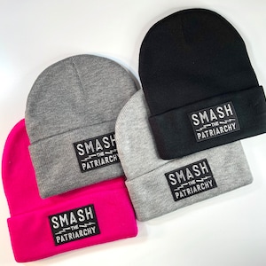 Puede incluir: Cuatro gorros de punto en diferentes colores con un parche blanco y negro que dice "Smash the Patriarchy".