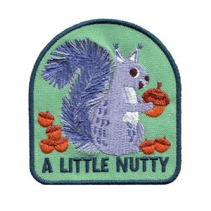May include: An embroidered patch with a light green background and a dark blue border. It features a light purple squirrel holding a brown acorn, surrounded by several other brown acorns. The text A LITTLE NUTTY is embroidered in dark blue.