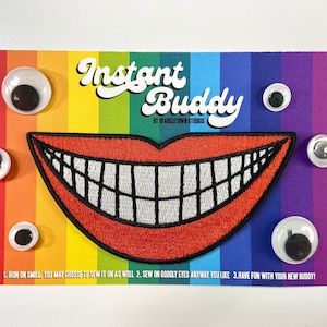 May include: A red felt smiling mouth with white teeth and black outline, surrounded by six googly eyes on a rainbow striped background. The text "Instant Buddy" is written in a retro font above the mouth. The text "By Sparkletown Studios" is written in a smaller font below the mouth. The text "1. Iron on smile, you may choose to sew it on as well. 2. Sew on googly eyes anyway you like. 3. Have fun with your new buddy!" is written at the bottom of the card.