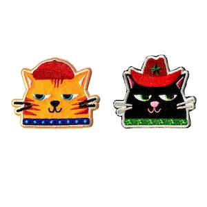May include: Two embroidered iron-on patches featuring cartoon cats. One cat is orange with a red hat and blue collar. The other cat is black with a red cowboy hat and green collar.