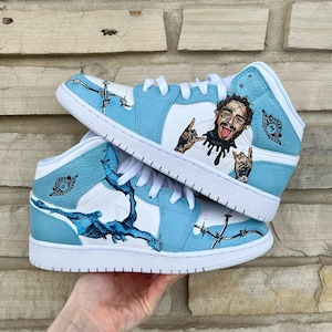 May include: A pair of custom-painted light blue and white high-top sneakers with a barbed wire design and a portrait of a man with a tongue sticking out. The sneakers have a white swoosh logo and the text "MONSTERCLO" on the side.