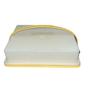 May include: A square, off-white plastic cake carrier with a yellow handle and base. The lid is clear, allowing a view of the contents. The handle arches over the top, and the base is a matching yellow. The carrier is designed for transporting and storing baked goods.