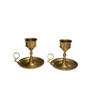 May include: Two brass candle holders. Each has a circular base, a central stem, and a cup-shaped top to hold a candle. A small handle is attached to the side of the base. The candle holders are made of shiny, polished brass.