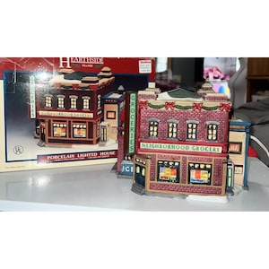 May include: A porcelain lighted house from the Earthside Village collection. The miniature building is a neighborhood grocery store with a red brick facade, lit windows, and a green awning. The sign reads "Neighborhood Grocery".