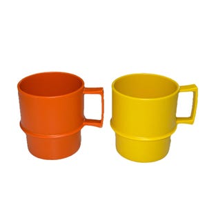May include: Two brightly colored mugs. One is orange, the other is yellow. Both mugs have a cylindrical body with a handle and a raised ring near the base. The mugs are made of a solid, opaque material.