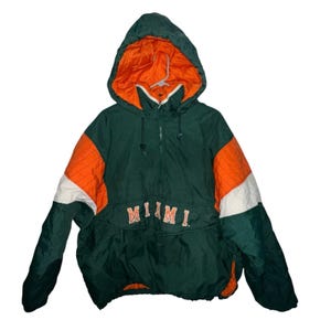 May include: A green, orange, and white hooded anorak jacket. The jacket has a half-zip closure and the word "MIAMI" is embroidered across the front in orange and white lettering. The hood is lined with orange fabric.