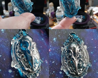 May include: A blue and silver pendant featuring a bird skull within an ornate, oval frame. The pendant has a detailed, gothic design with a glossy finish. The background is a galaxy pattern.