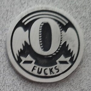 May include: A silver-colored metal coin with a black design. The coin features a large letter "O" with a tire tread pattern inside. The word "FUCS" is printed below the letter.