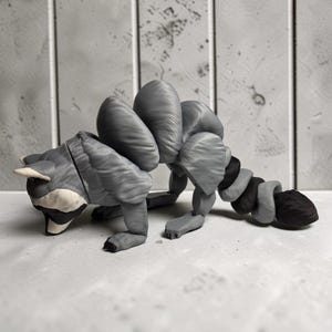 May include: A gray and black articulated raccoon figurine featuring a segmented back and a flexible, multi-part tail. The figure has a distinct mask pattern around the eyes and is constructed with visible joints that allow for movement and posing.