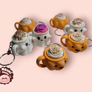 May include: Keychains featuring miniature coffee cups and ghost-shaped mugs with whipped cream toppings. The cups are orange and white, some with smiling faces. A purple-topped cup is also present. Each keychain has a silver ring.