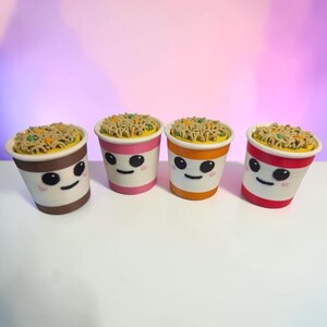 May include: Four small cylindrical containers featuring a smiling face with black eyes and a thin mouth. Each container is topped with a textured representation of noodles, including small green and orange pieces, in brown, pink, orange, and red.