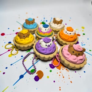 May include: A collection of seven bear-shaped erasers designed to look like miniature tarts with colorful frosting. Each bear has a distinct color, including yellow, blue, white, orange, purple, and pink. Some include attached ball chains.