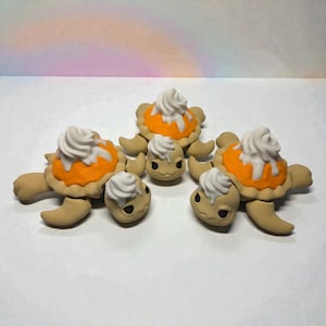 May include: Five whimsical turtle figurines. Each turtle has a tan body and limbs, with an orange pie shell and white whipped cream topping on its back. The turtles are arranged on a white surface.