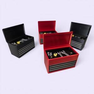 May include: Four miniature tool chests with open lids, two in red and two in black. Each chest features horizontal gray drawer fronts and contains small silver-colored wrenches, nuts, and yellow components arranged inside the open top compartment.