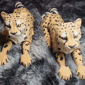 May include: Two tan cheetah figurines with black spots are lying down on a gray, textured surface. The cheetahs have detailed facial features and articulated limbs. The image also includes a circular logo with a horse illustration.