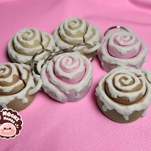 May include: Six cinnamon roll keychains with white icing and silver key rings. The rolls are in shades of brown, tan, and pink, set against a pink background. The keychains are a fun, food-themed accessory.
