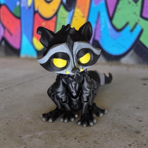 May include: A 3D-printed raccoon figurine with a black body, gray face mask, and yellow eyes. The raccoon is posed in a sitting position. The background features colorful graffiti art.
