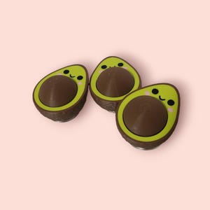 May include: Three avocado-shaped push pop fidget toys with brown pits and green avocado skin. Each avocado has a cute cartoon face with pink cheeks and a smile.