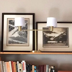 May include: A gold metal double-arm lamp with two white lampshades. The lamp is sitting on a wooden shelf with books and a small wooden figurine.