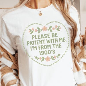 May include: White long-sleeve t-shirt with a floral heart design and the text "PLEASE BE PATIENT WITH ME, I'M FROM THE 1900'S" in green. The shirt is a casual style, suitable for everyday wear. The design is a vintage-inspired graphic.