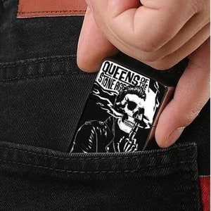 Può includere: Un accendino bianco e nero con la scritta "Queens of the Stone Age" stampata su di esso. L'accendino presenta una grafica di una persona con un viso di teschio che indossa una giacca di pelle.
