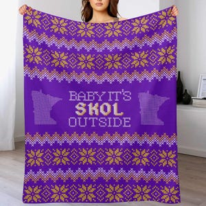 Puede incluir: Manta morada con un patrón repetido de copos de nieve dorados y blancos y formas geométricas. La manta tiene la frase "BABY IT'S SKOL OUTSIDE" en blanco y dorado, con un contorno de Minnesota a cada lado.